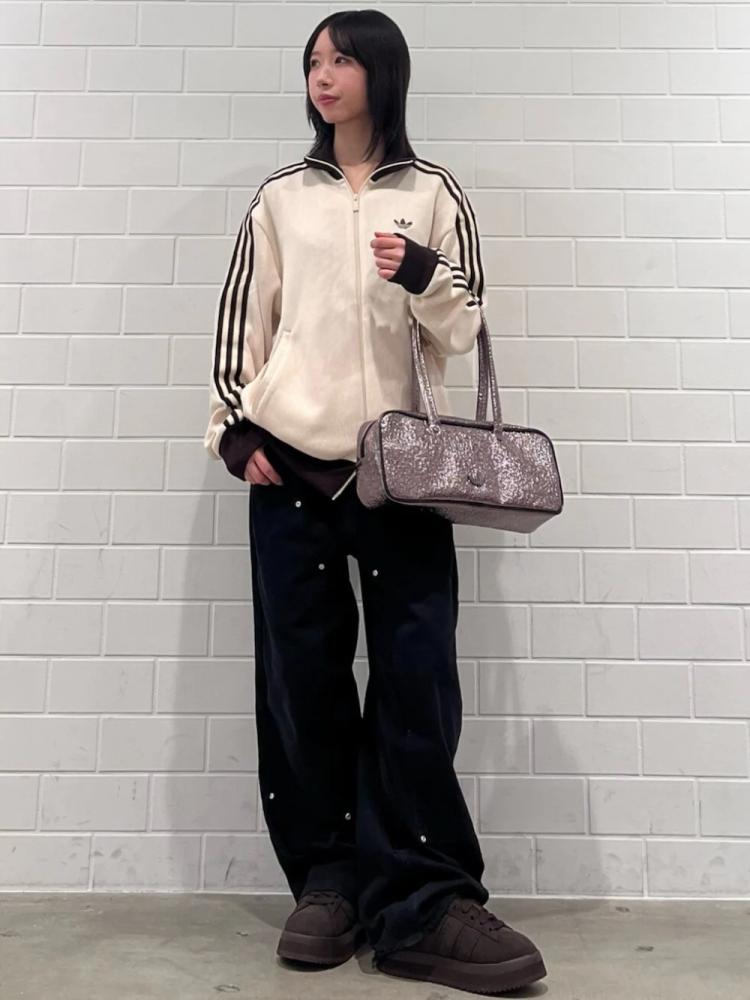 A young woman wearing a beige hooded jacket, black pants, and dark shoes stands in front of a white tiled wall, holding a silver handbag.
