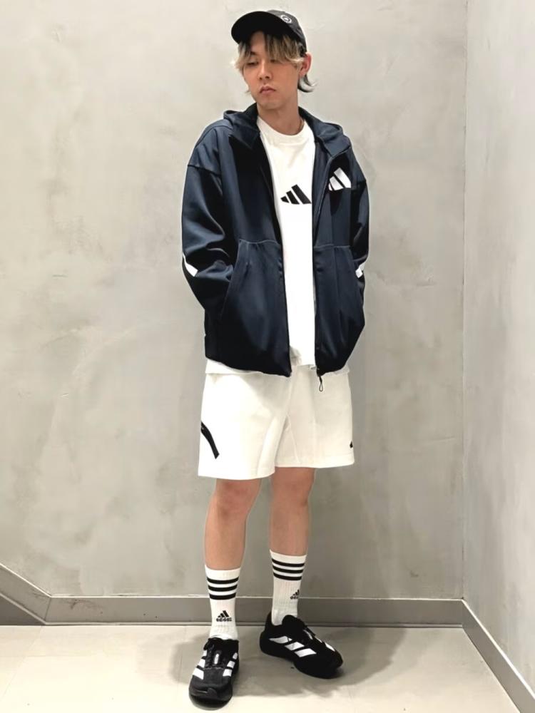 A young person wearing a navy blue hooded jacket, white t-shirt, and white shorts stands against a plain white wall.