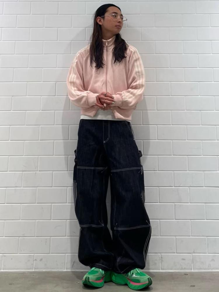 A person wearing a pink sweater, black pants, and green shoes stands in front of a white brick wall.