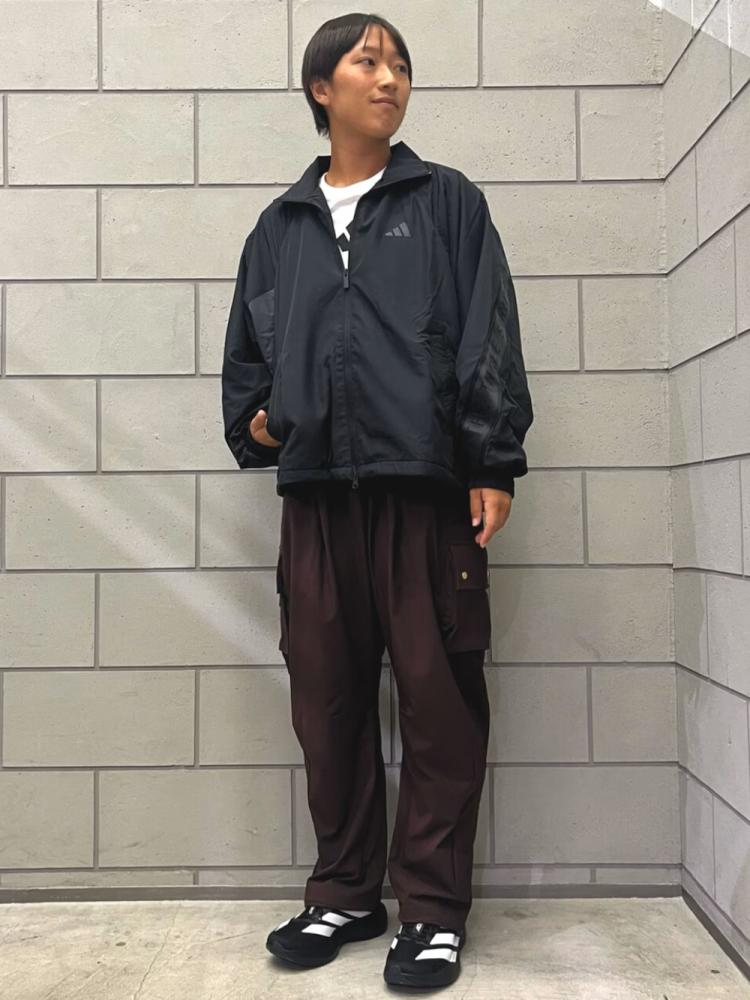 A young person wearing a black jacket and dark pants stands in front of a gray brick wall.