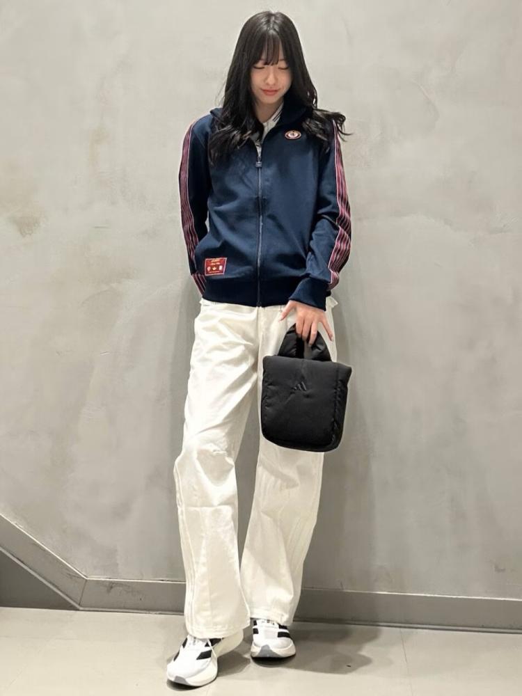 A young woman wearing a navy blue jacket, white pants, and sneakers stands in front of a plain gray wall, holding a black handbag.