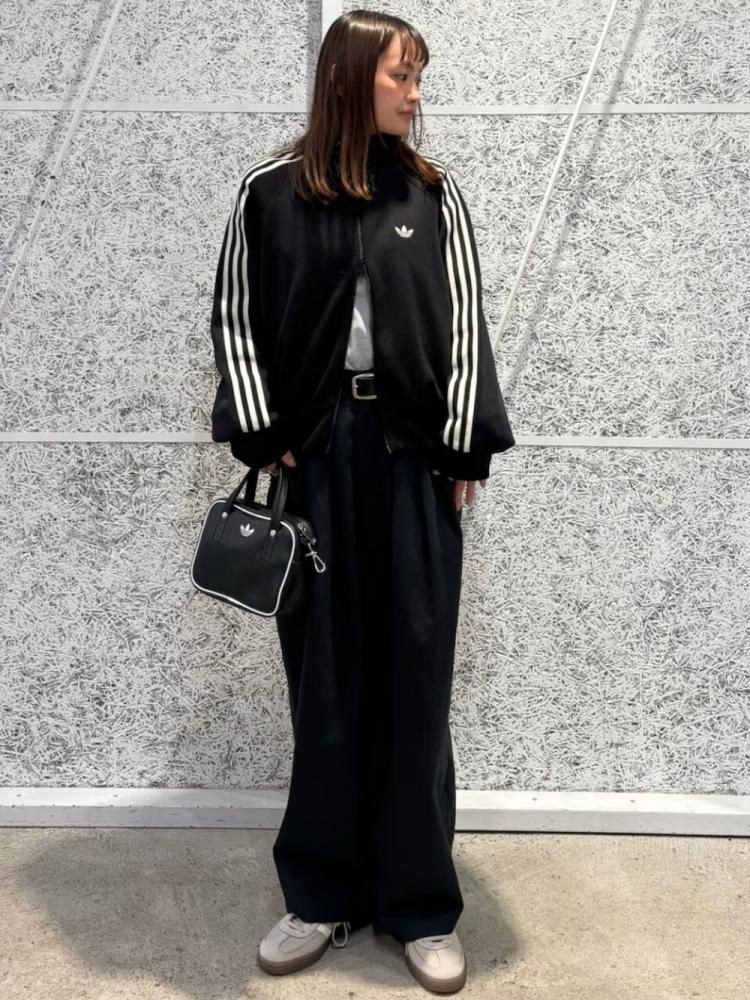 A young woman wearing a black Adidas tracksuit stands in front of a gray concrete wall, holding a black handbag.
