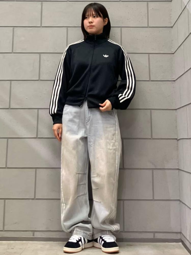 A young person wearing a black Adidas jacket and gray sweatpants stands in front of a gray concrete wall.