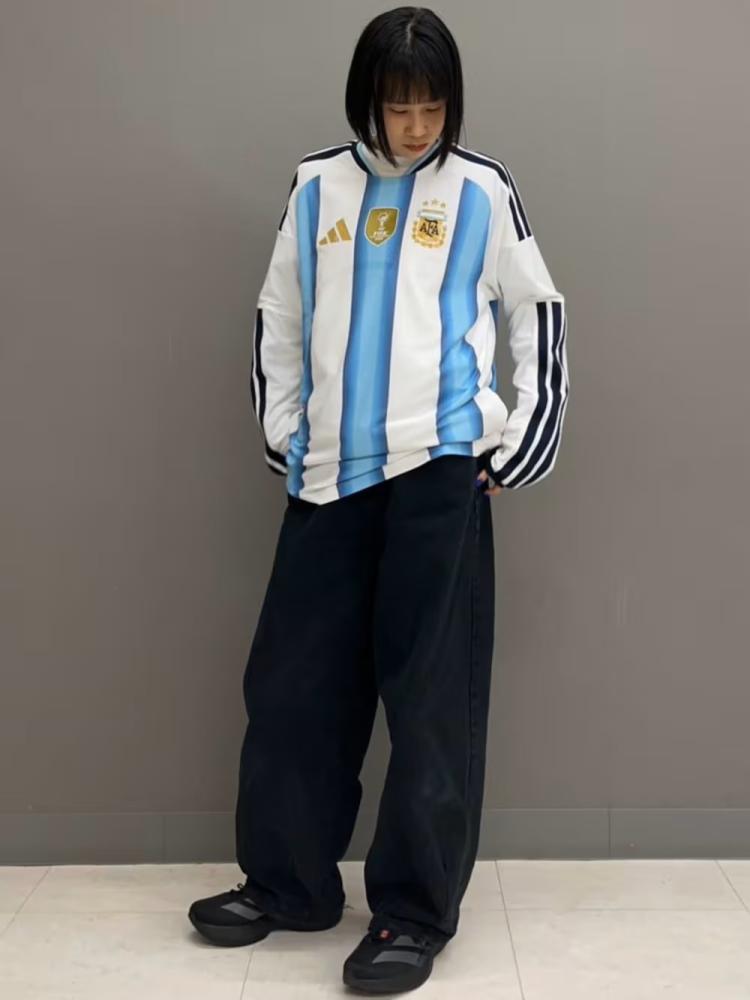 A young person wearing an Argentinian national team jersey and black pants stands in front of a plain gray background.