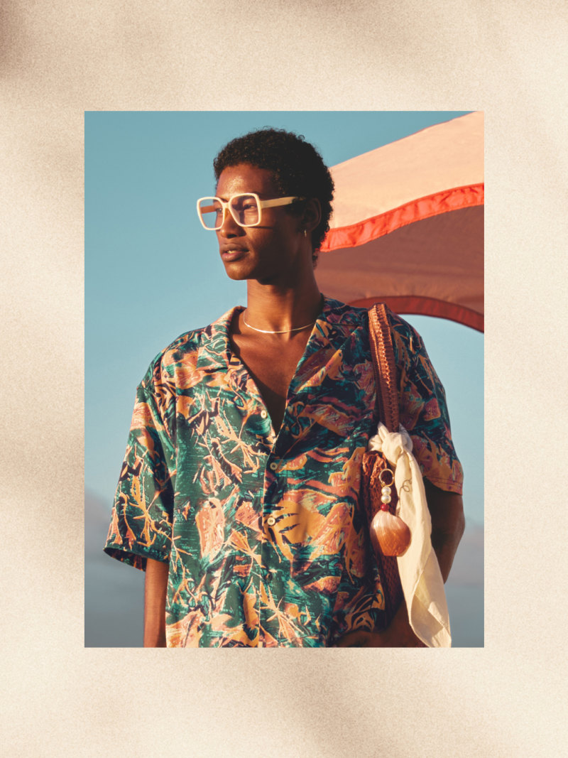 Un hombre con una camisa y gafas coloridas y estampadas se coloca de pie contra un cielo azul brillante, sosteniendo un paño o una bufanda blanca.