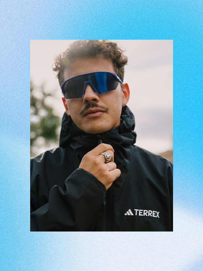 Una persona con chaqueta negra y gafas de sol se coloca de pie en un entorno exterior con un cielo azul de fondo.
