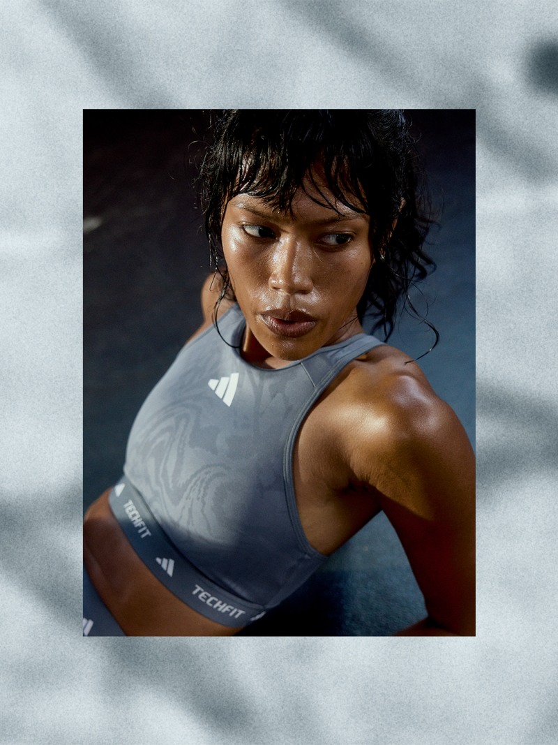 A person with dark hair and a determined expression, wearing a gray athletic top, stands against a blurred background.