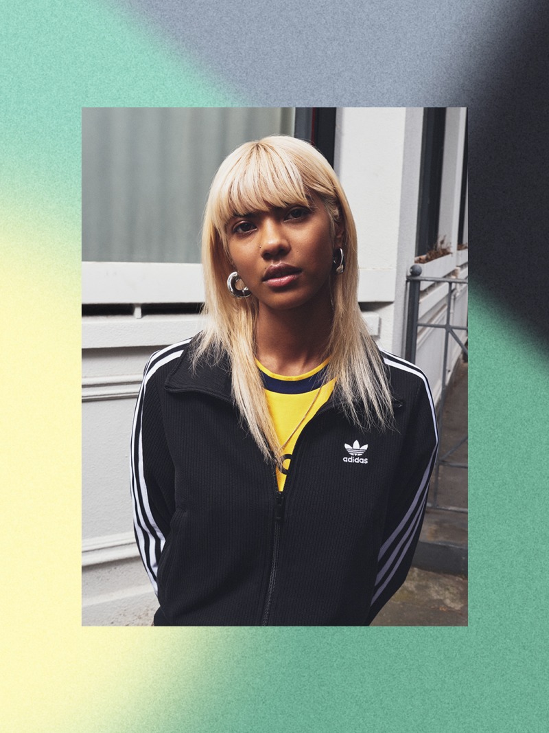 Mladá žena s blonďavými vlasmi v čiernej bunde adidas stojí pred sivým pozadím.