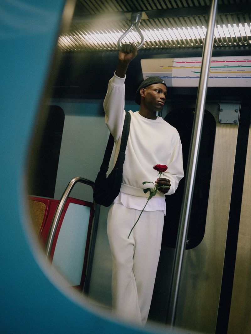 A person in a white uniform stands in a subway car, holding onto a pole and holding a red object, with a blue and white background visible.