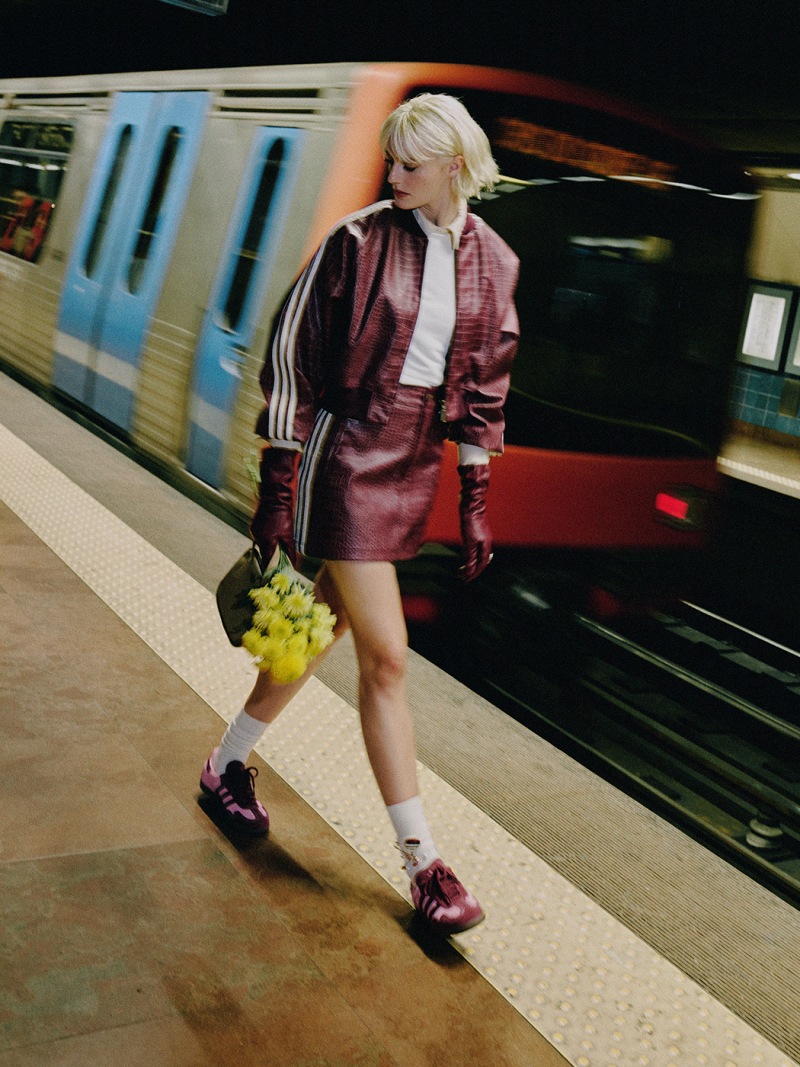 A young woman in a pink leather jacket and shorts stands on a subway platform, holding a bunch of grapes, as a train rushes by in the background.