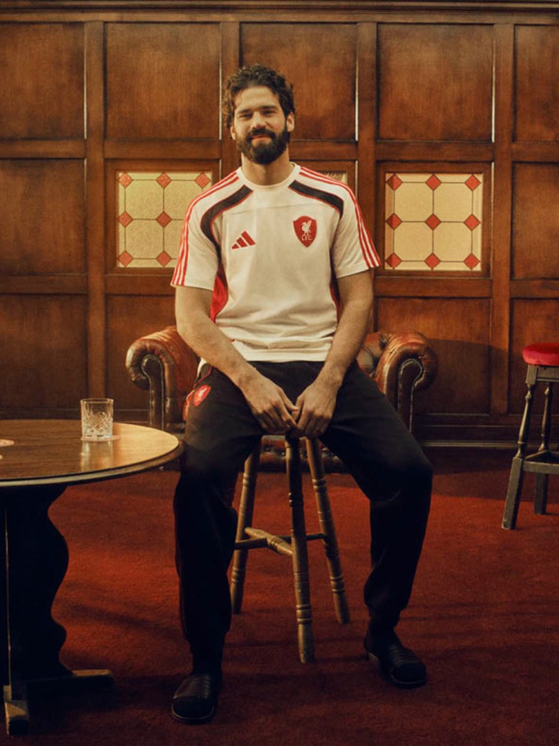 A man with a beard wearing a white and red sports jersey is seated in a wooden chair in a room with ornate wooden paneling and patterned tile flooring.