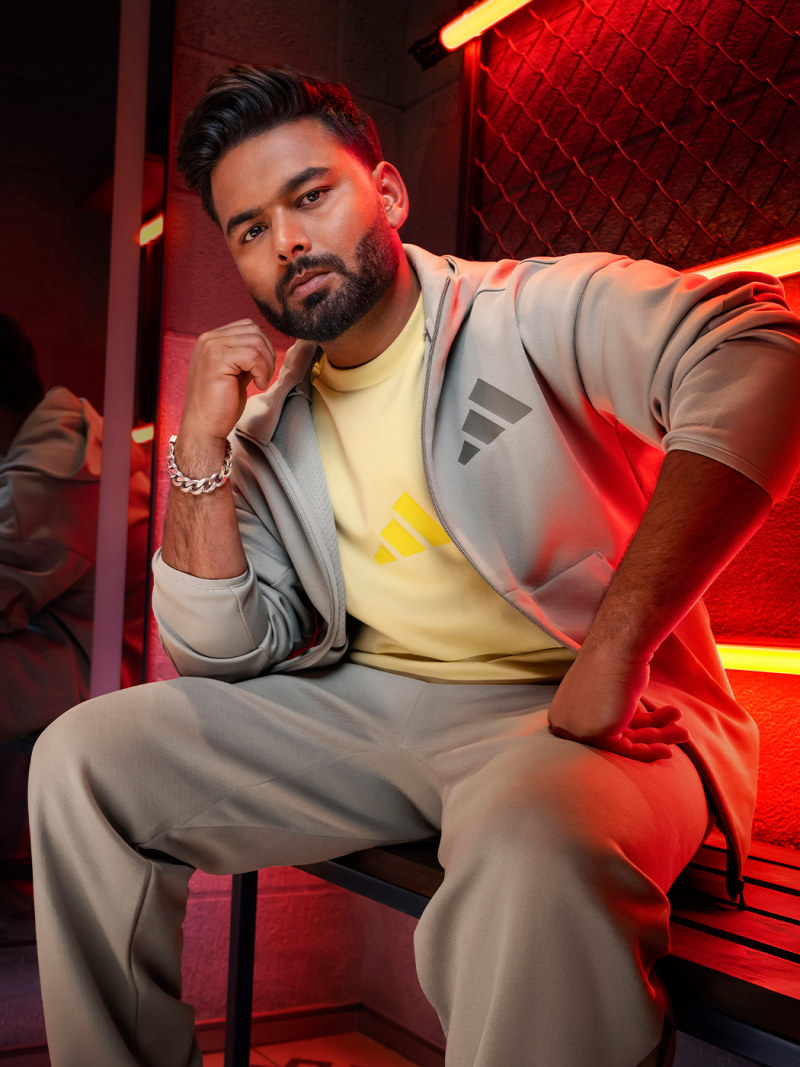 A young man with dark hair and a serious expression sits in a gray outfit against a vibrant green background, with red lighting casting an intense glow on his face.