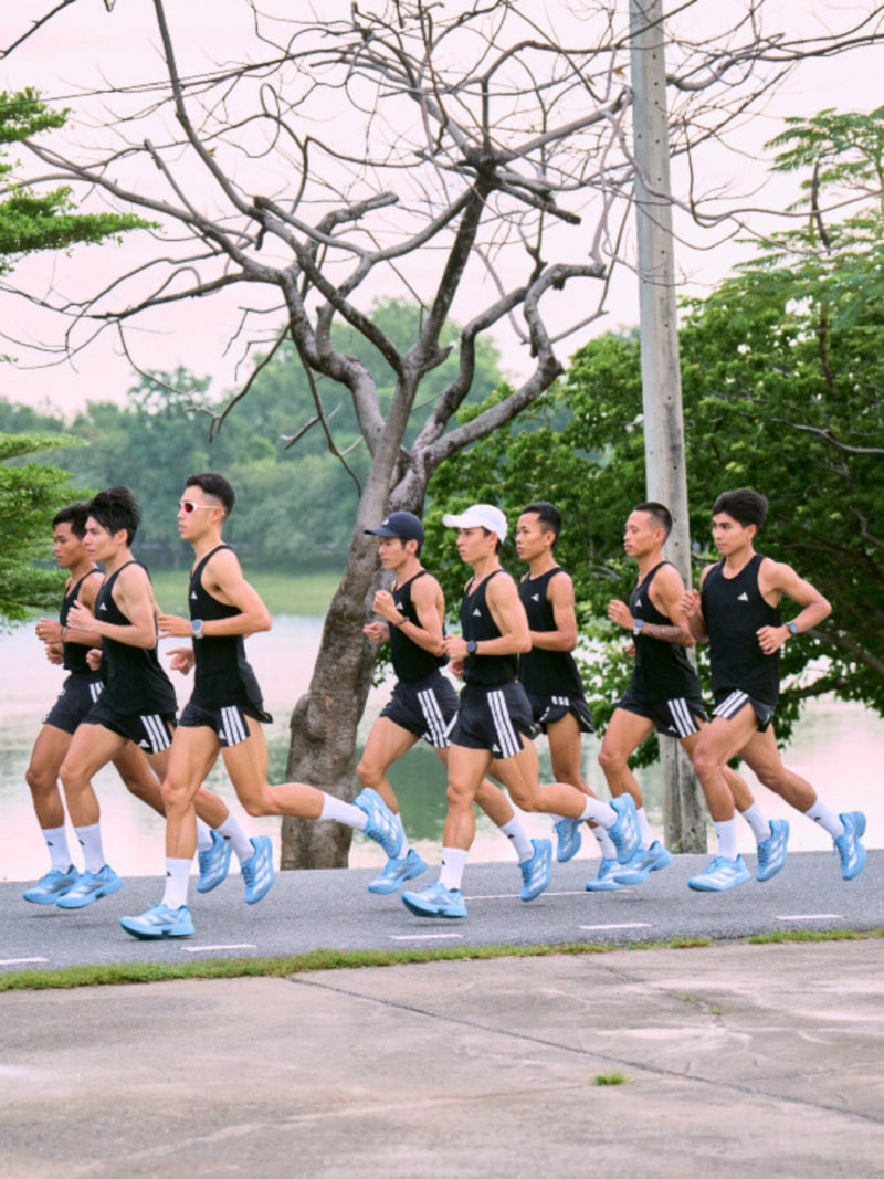 A group of runners in athletic gear are jogging along a path surrounded by lush greenery and a bare-branched tree in the foreground.
