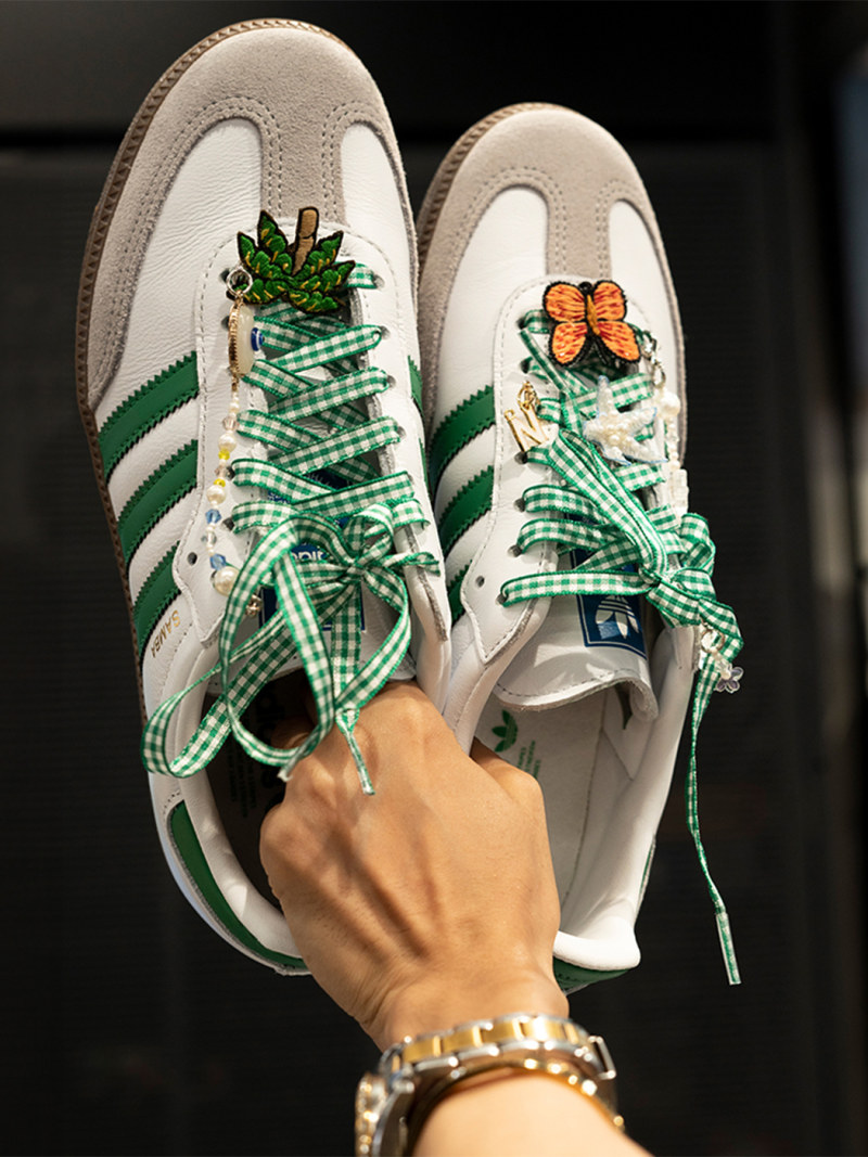 A pair of white and green sneakers with decorative laces and embroidered floral and butterfly designs, being held by a hand adorned with bracelets.