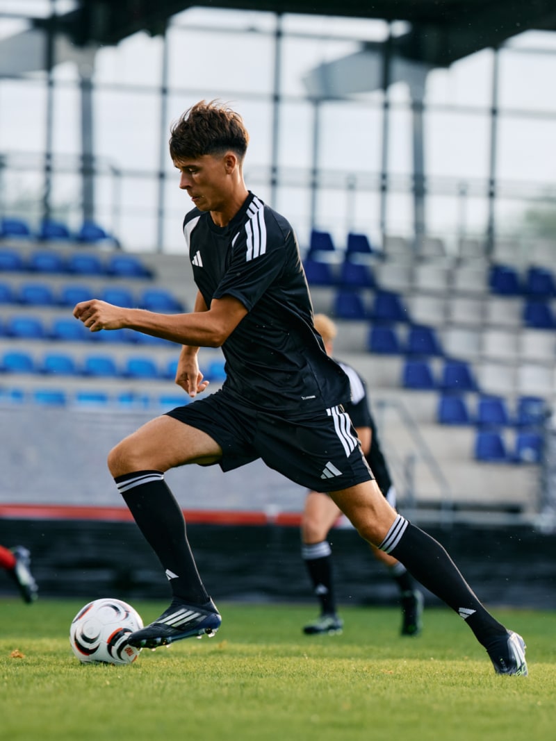 A young soccer player in a black uniform is dribbling a ball on a soccer field with stadium seating visible in the background.