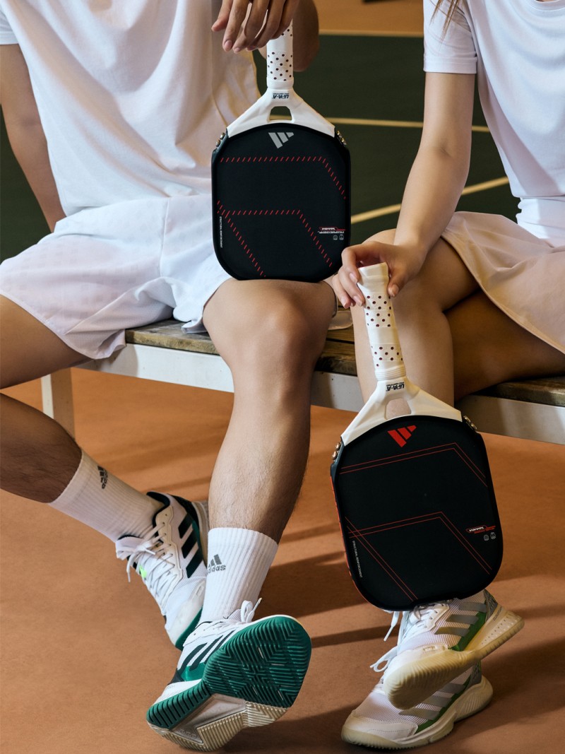 The image shows a person's lower body sitting on a wooden floor, wearing white socks and sneakers, with two small black bags or pouches placed on the floor next to them.