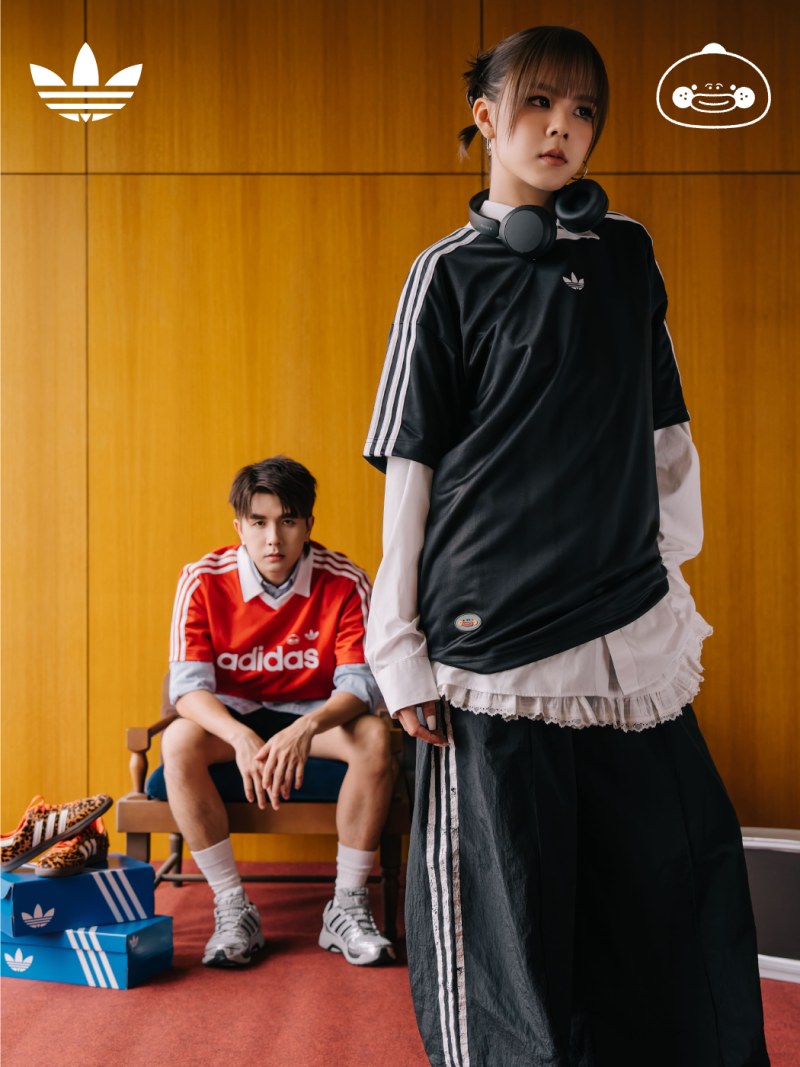 Two young individuals, one standing and one sitting, wearing Adidas sportswear against a wooden background with Adidas and gaming console logos.