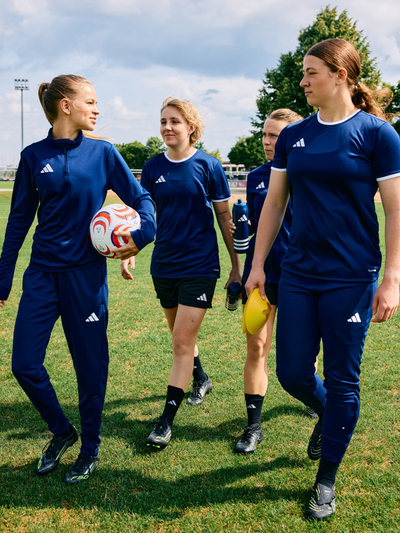 A group of young female soccer players in blue uniforms walking together on a grassy field, with trees and a cloudy sky visible in the background.