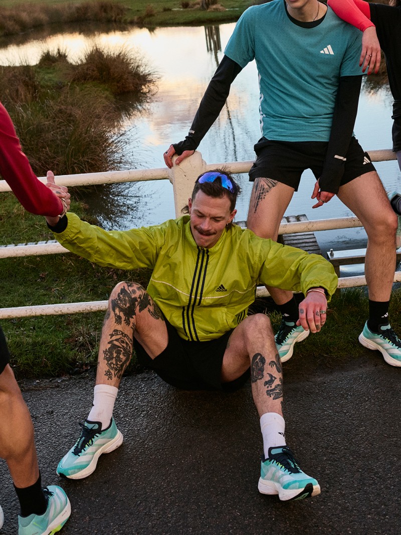 A smiling man in a bright yellow jacket sits on the ground surrounded by other people in athletic clothing, with a body of water and natural scenery visible in the background.