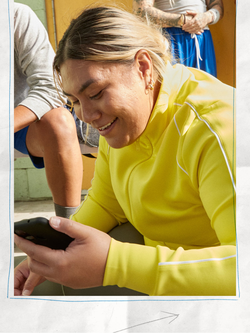 A young woman in a bright yellow sweater is smiling and using a smartphone while sitting in a colorful, cluttered room.