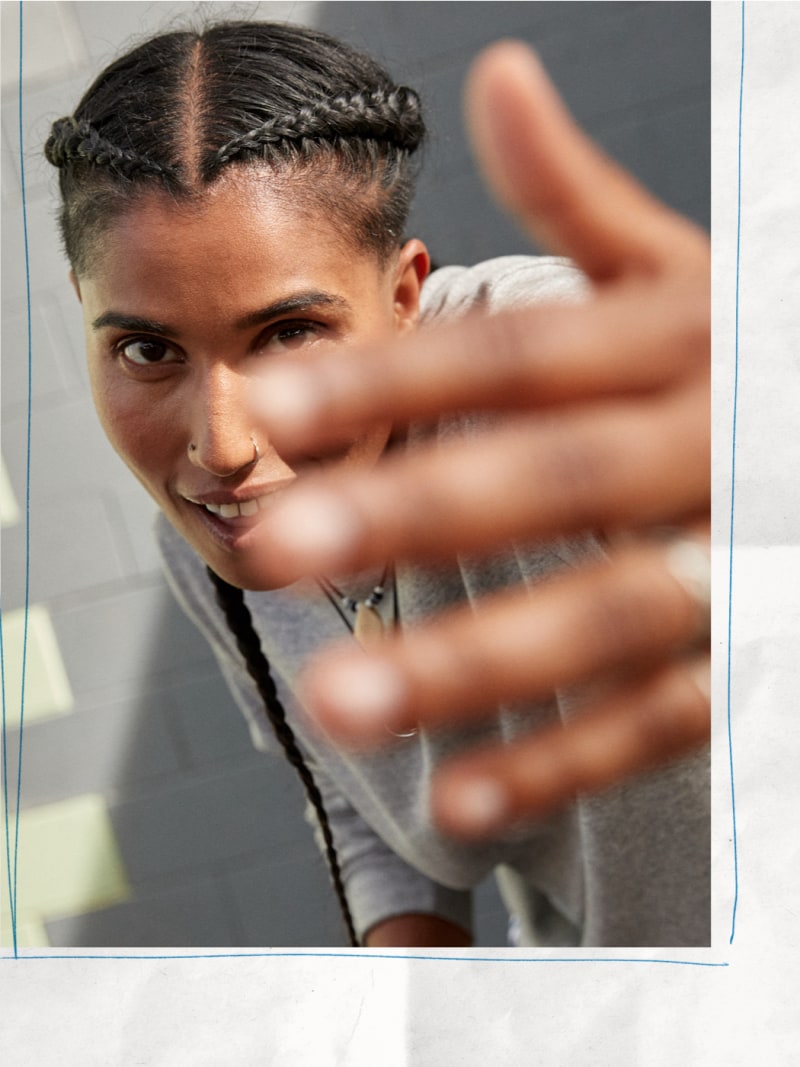 The image shows a person with braided hair making a hand gesture in the foreground, with a blurred background.