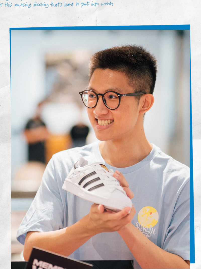 A young man wearing glasses and a white t-shirt is smiling and holding a pair of white sneakers in the foreground, with a blurred background of what appears to be an indoor setting.