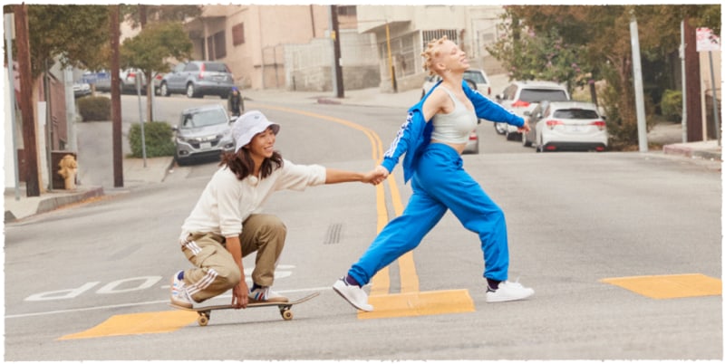 An image with two people crossing the road holding hands. The girl walks straight and the boy rides a skateboard
