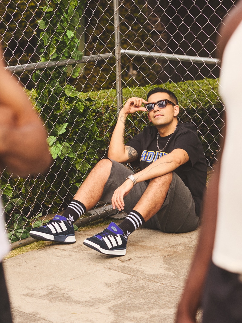 A person wearing a black shirt, sunglasses, and sneakers is sitting on the ground in front of a chain-link fence with greenery in the background.