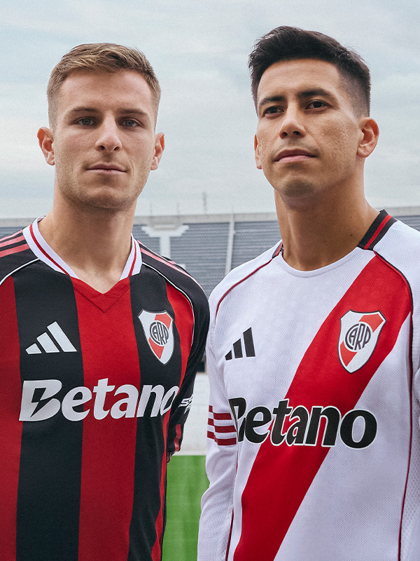 Two young men wearing soccer jerseys with the team name "Betano" stand in front of a stadium or sports facility.