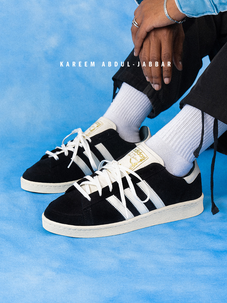 The image shows a pair of black and white sneakers with the name "Kareem Abdul-Jabbar" printed on them, set against a bright blue sky background.