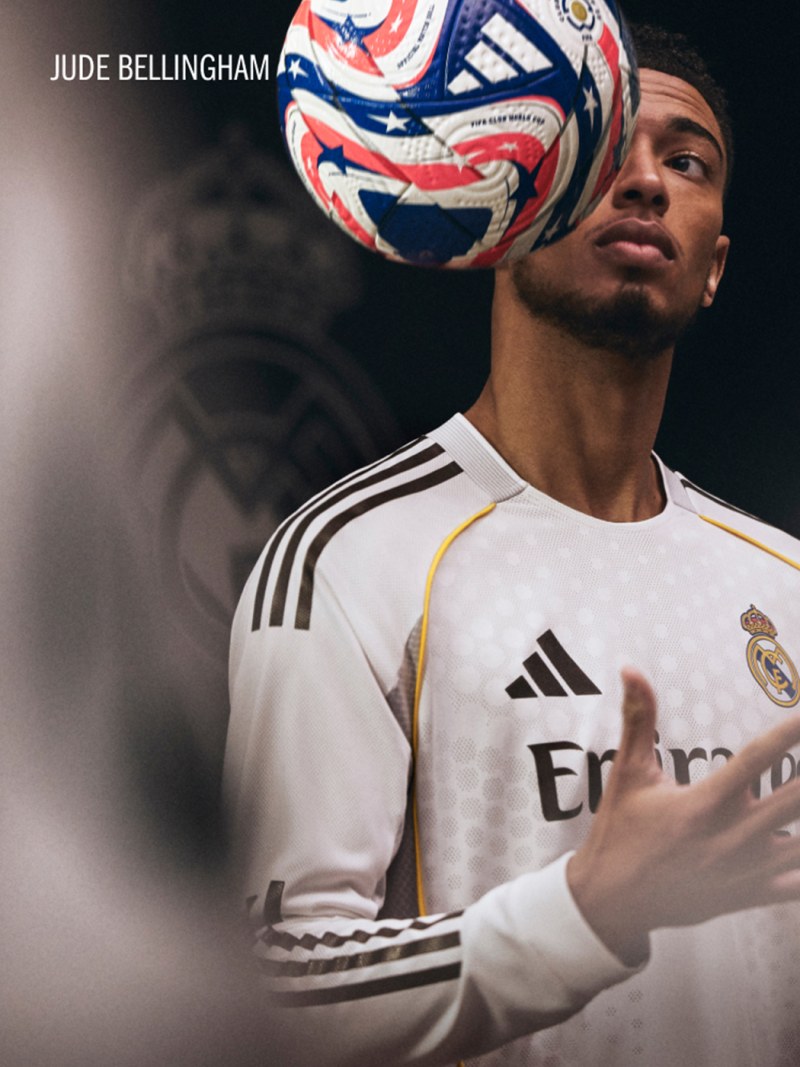 A young man wearing a white soccer jersey with the Real Madrid logo stands holding a colorful soccer ball, with a dark background behind him.