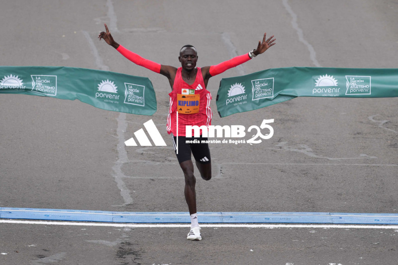 A runner in a red and white uniform with arms raised in celebration crosses the finish line in front of a backdrop of event banners and signage.