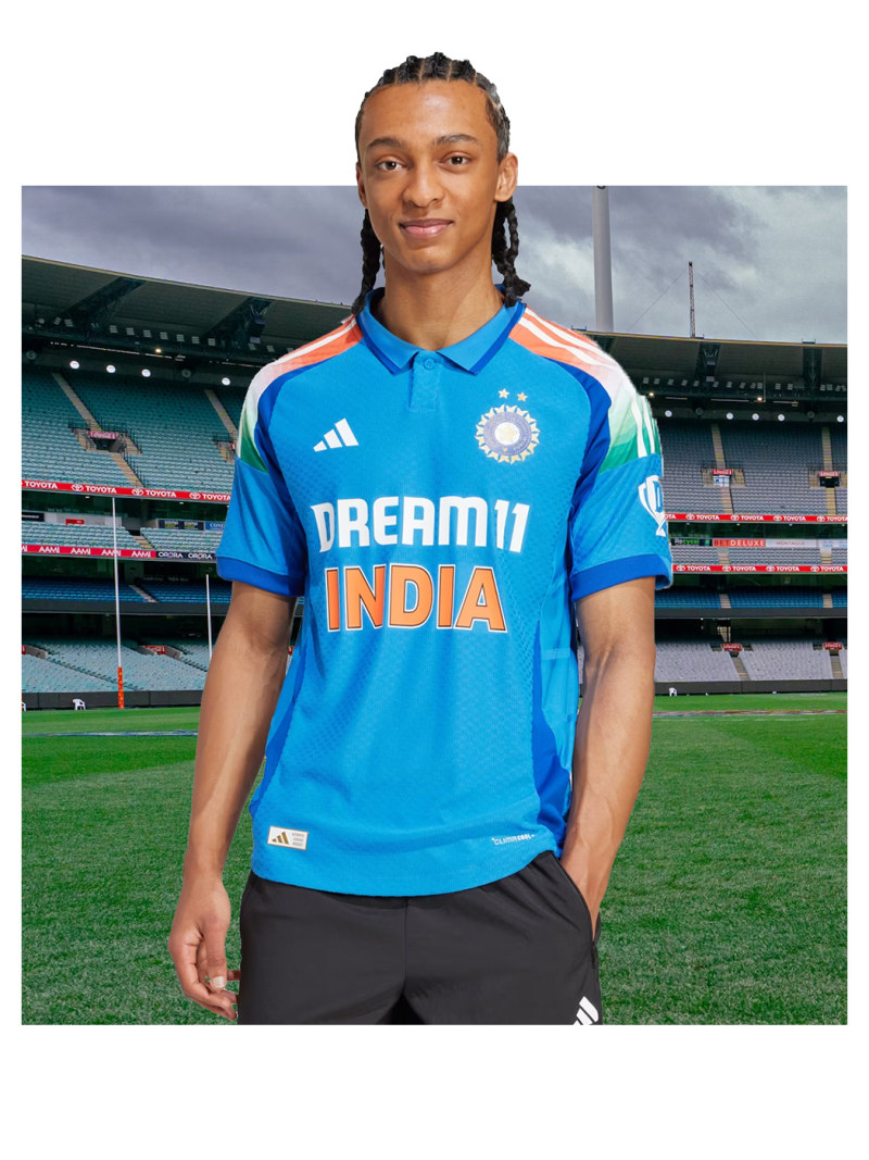 A young man wearing a blue and orange cricket jersey stands on a cricket field with a stadium in the background.