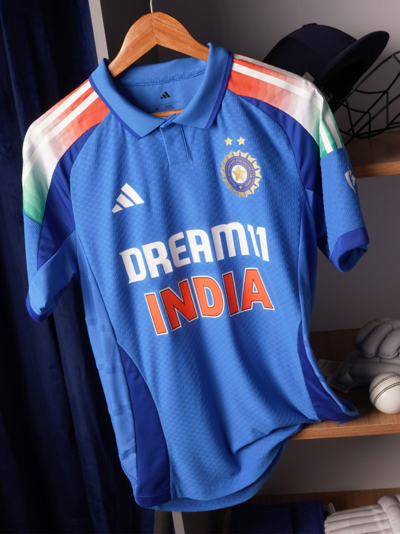 A blue cricket jersey with the text "DREAM INDIA" is displayed on a wooden hanger against a dark background.