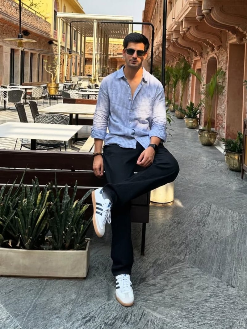 A well-dressed man sits on a bench in an outdoor setting with ornate architectural features and potted plants in the background.