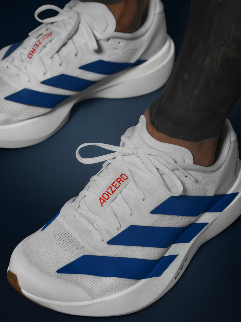 The image shows a pair of white and blue athletic shoes with the Adidas logo prominently displayed on the side. The shoes are set against a dark blue background.