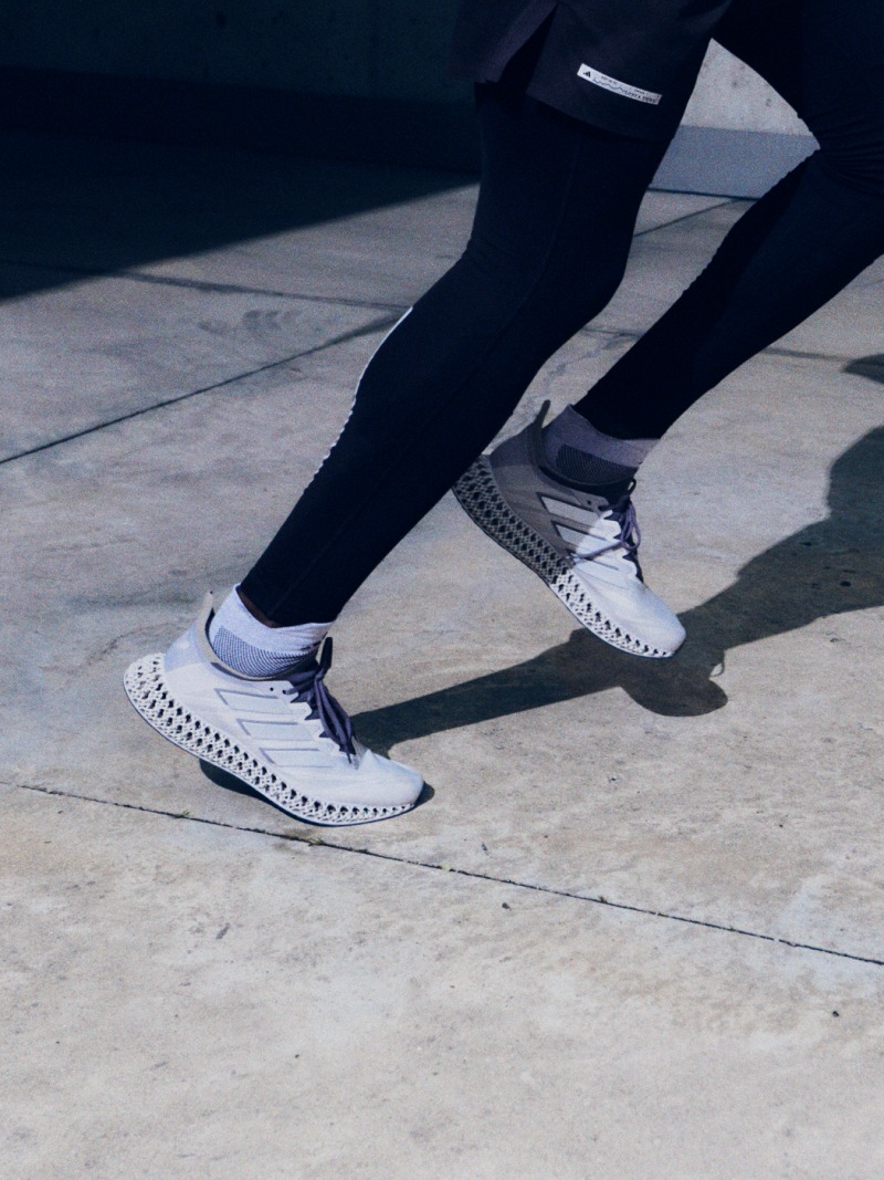 The image shows a close-up view of a white athletic shoe with a textured, patterned sole and upper. The background is dark, creating a dramatic contrast that highlights the intricate design and structure of the shoe.