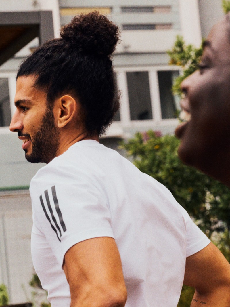 Two men, one wearing a white shirt and the other wearing a dark shirt, are standing in a garden setting with plants and flowers visible in the background.