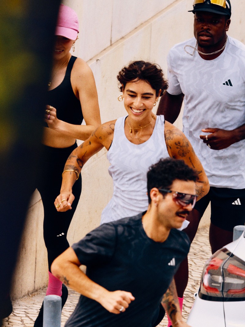 A group of young people, including a woman in a black tank top, a man in a white shirt, and another man in a black shirt, are walking together in an urban setting with a car in the background.