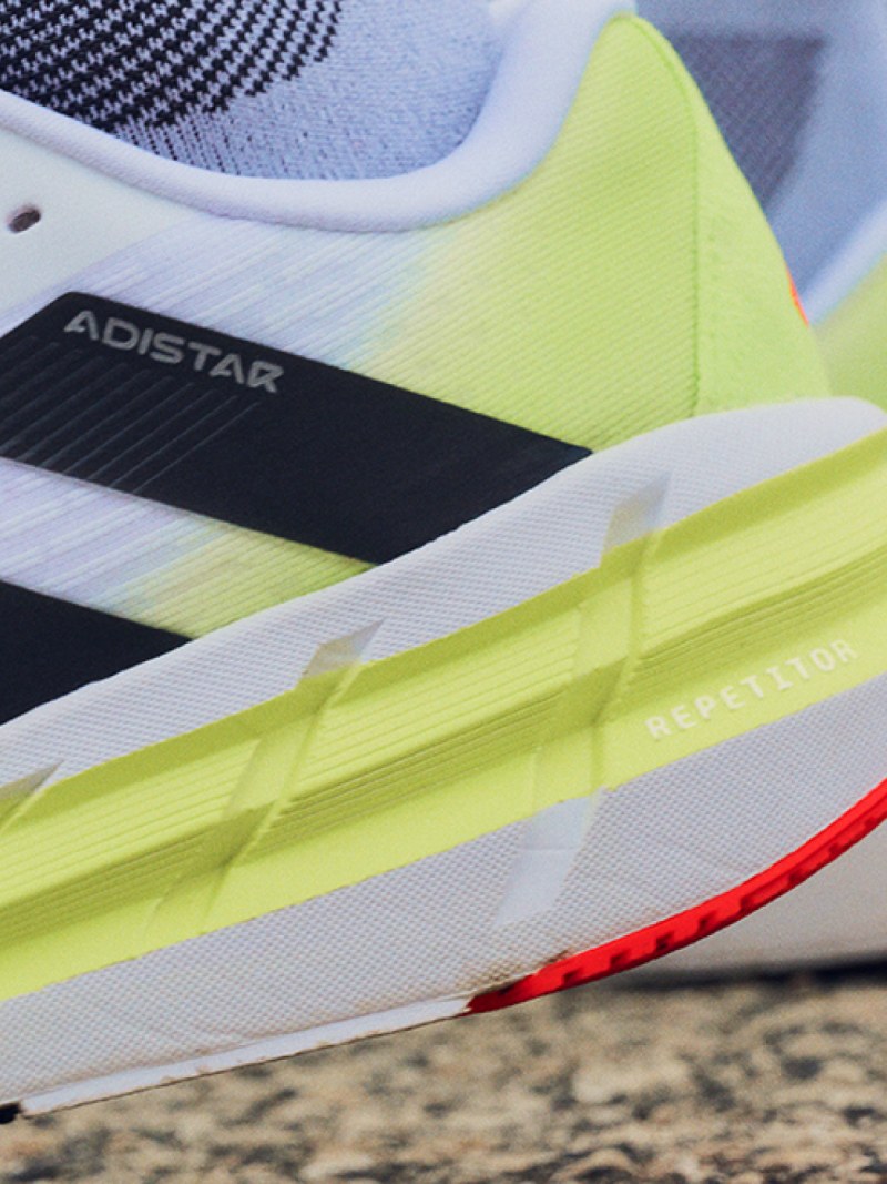 The image shows a close-up view of a running shoe, featuring a bright yellow and white design with black accents. The shoe appears to be resting on a textured surface, possibly a concrete or asphalt background.