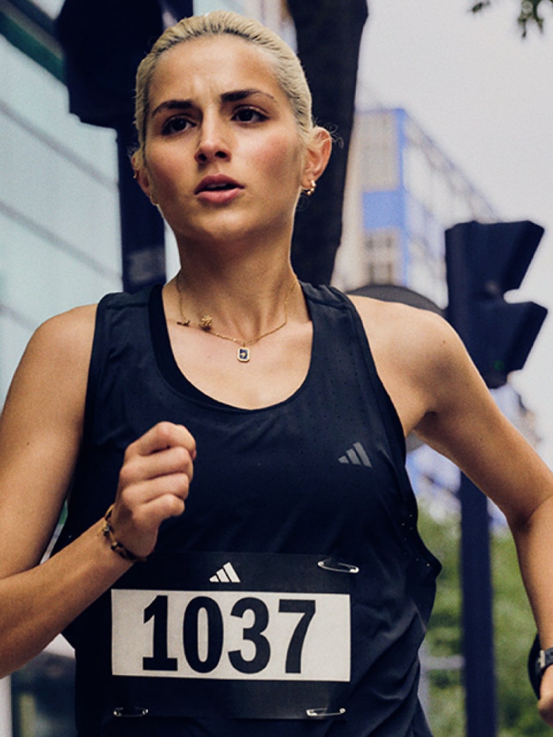 A young woman wearing a black athletic top and shorts, with a race bib number 1037 pinned to her shirt, is running on a city street with buildings in the background.