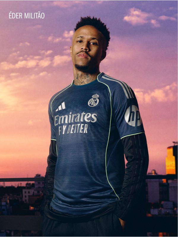 The image shows a man wearing a navy blue soccer jersey with the Emirates logo, standing against a vibrant sunset sky with clouds.