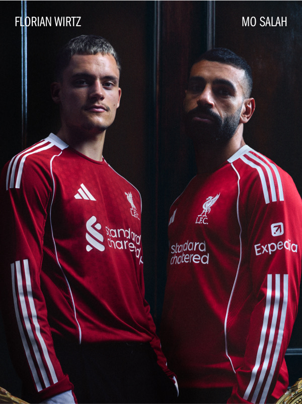 Two professional soccer players wearing Liverpool FC jerseys stand in a dark setting, their expressions focused and determined.