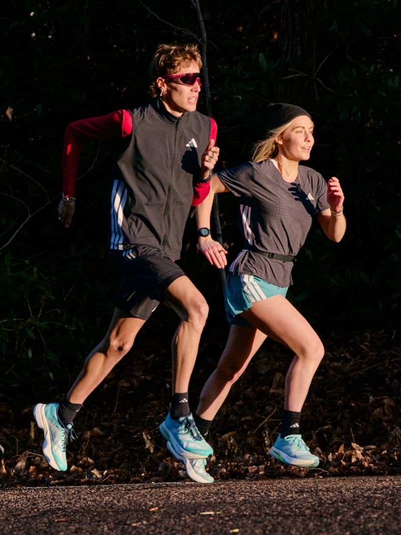 A man and a woman run at dusk on a country road, towards the sunlight, wearing the new Adizero Boston 13.
