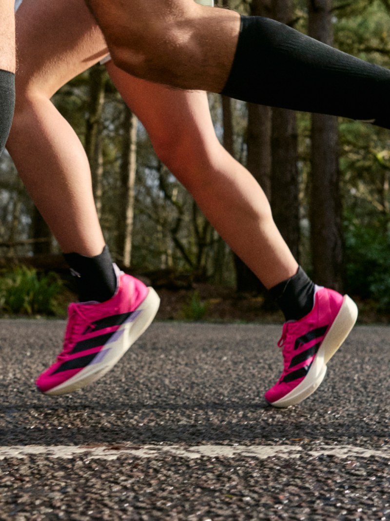 The image shows a person's lower body and legs, wearing bright pink athletic shoes, running on a paved path surrounded by a forested area.