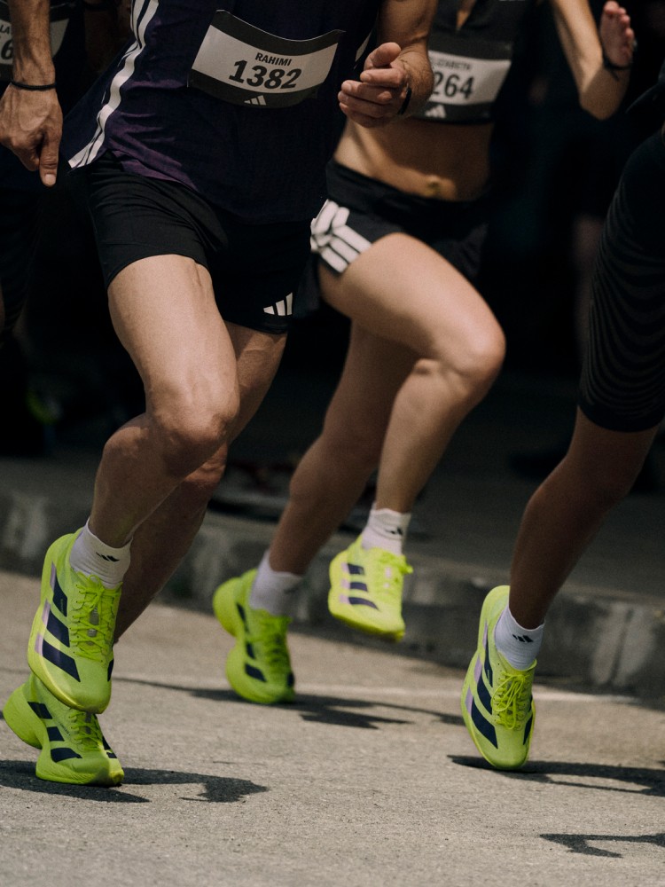 The image shows the lower bodies and legs of runners wearing bright green and black athletic shoes, with race numbers visible, suggesting a running event or competition taking place in a dark environment.