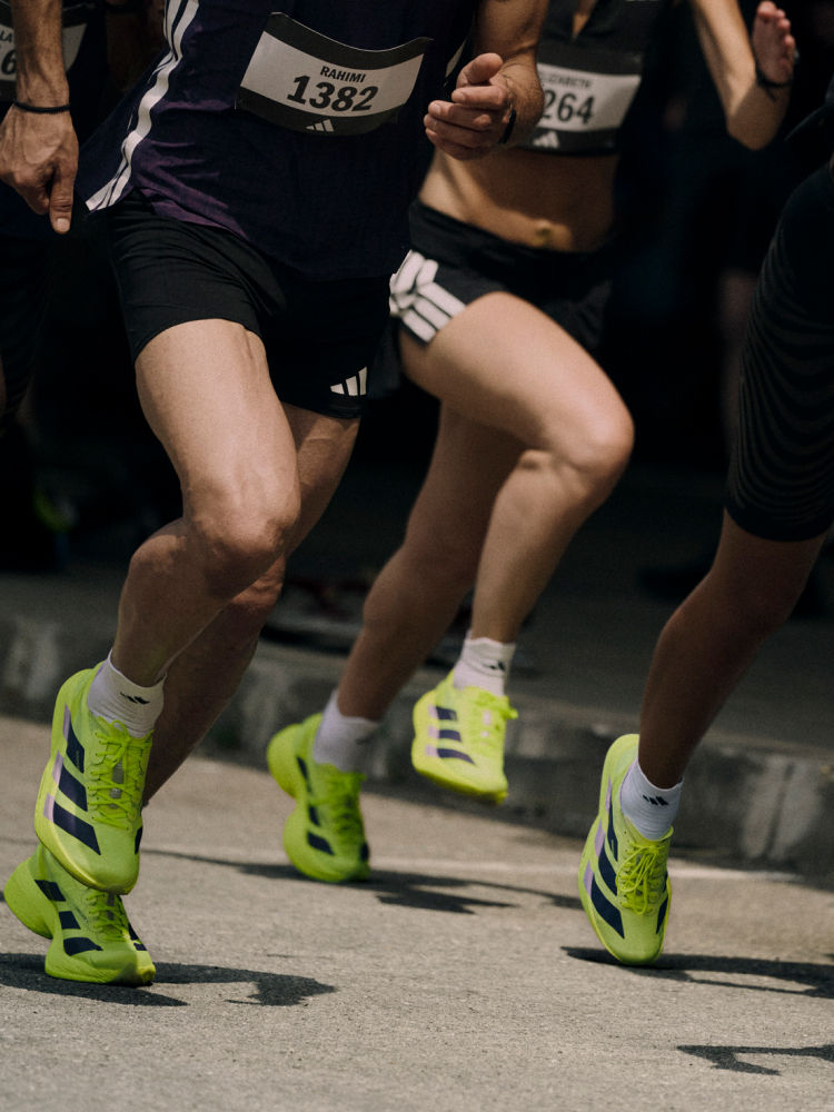 The image shows the lower bodies and legs of several runners wearing bright green and black athletic shoes, with race numbers visible, suggesting a running event or competition taking place in a dark environment.