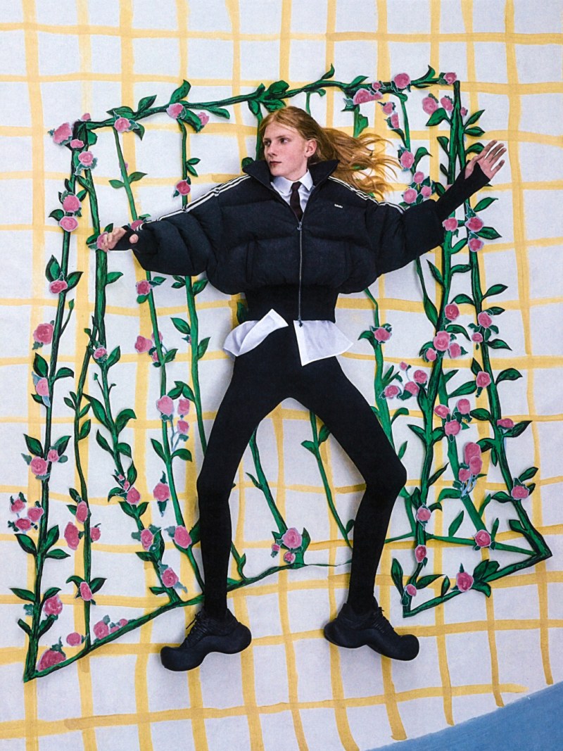 A person in a black outfit is standing with arms outstretched in a floral frame against a yellow and white checkered background.