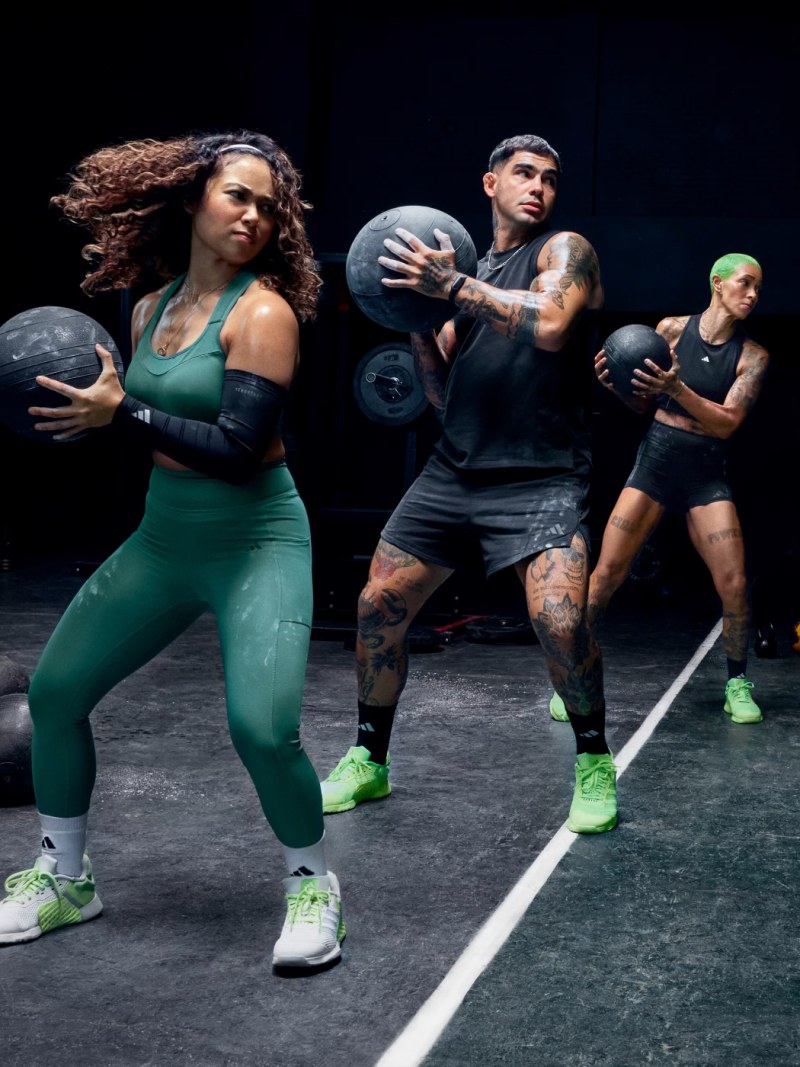Two women and a man wearing the adidas Dropset 3 strength training shoes tosses a ball in a dimly lit gym setting.