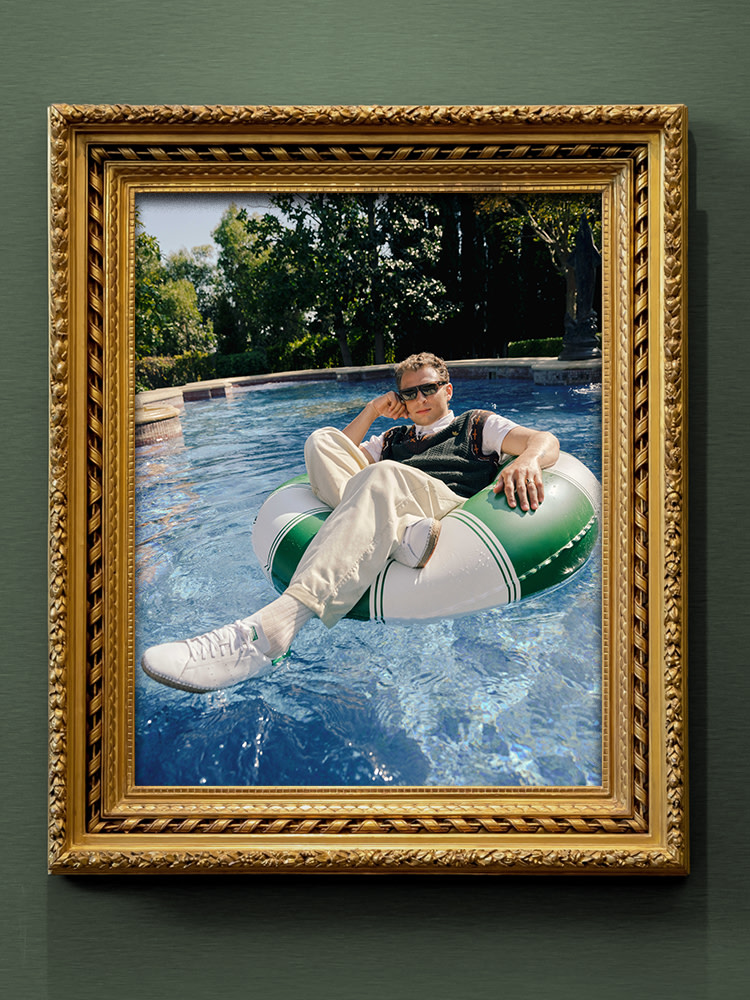 A man wearing the adidas X Edison Chen CLOT Stan Smith Collection, including sunglasses, white Stan Smith sneakers and a green knitted sweater vest lounges on a green and white pool float in a swimming pool, shown inside an ornate gold frame.